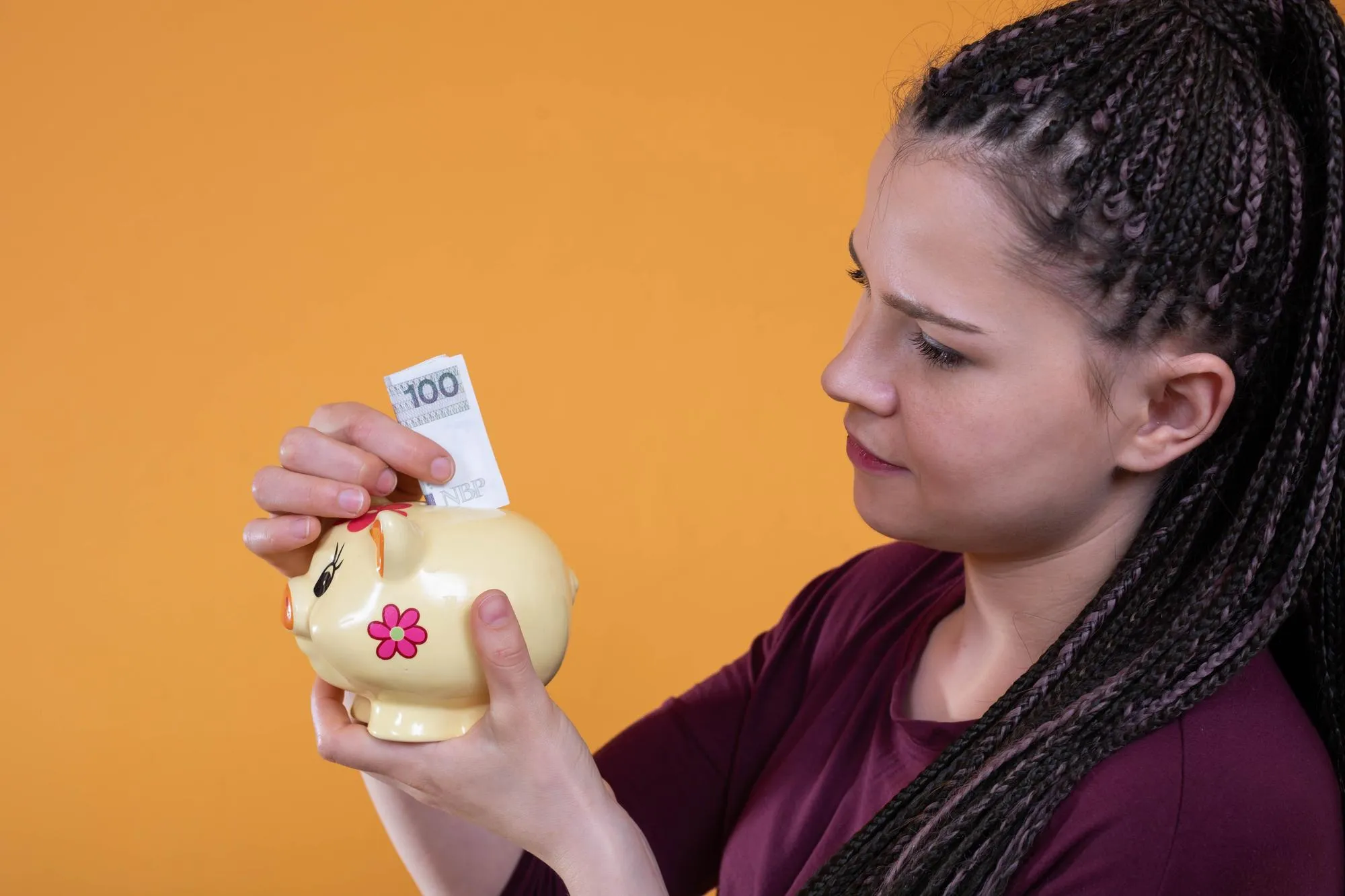 A young girl learns to save and puts a hundred Polish zloty banknote in her porcelain piggy bank Whole isolated on an orange background