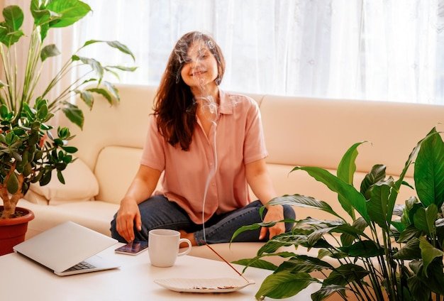 A young woman is resting from work sitting on the sofa and inhaling the fragrance of incense Blurry