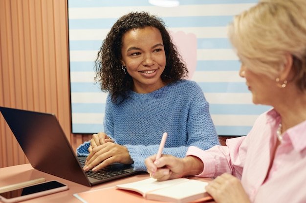 Young woman using laptop consulting senior colleague sitting at table in coworking
