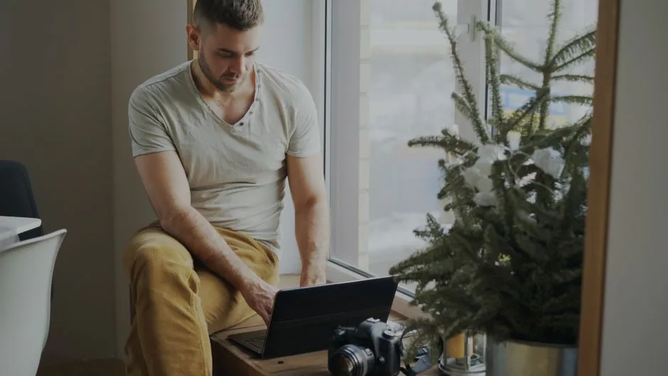 Casual man using laptop indoors by a window, with camera and plant nearby.