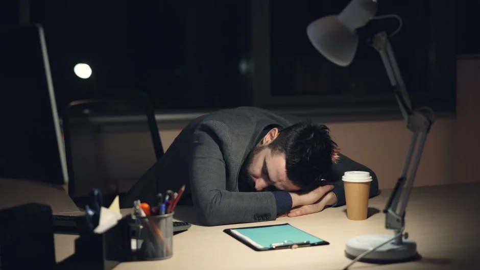 Man in office suit sleeping on desk with coffee and tablet nearby, symbolizing work fatigue.
