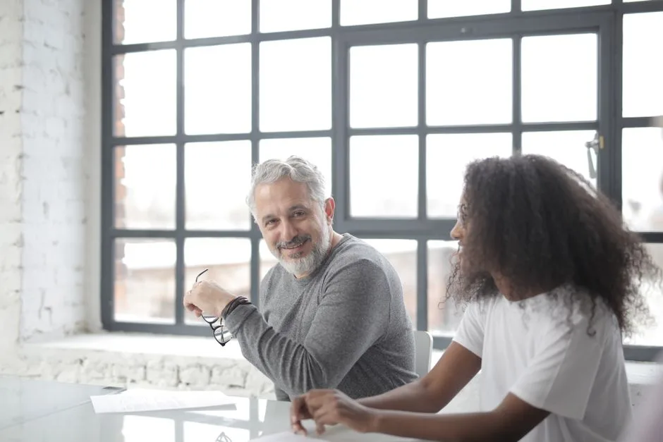 Happy aged gray haired coach in casual wear looking at camera while supporting young African American coworker in office boardroom