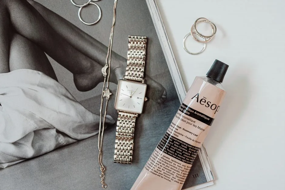 Stylish flat lay featuring a wristwatch, necklace, rings, and Aesop hand balm.