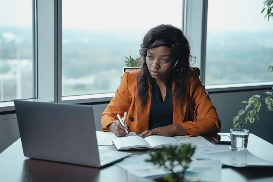A businesswoman in a modern office intently working on a laptop and writing in a notebook.