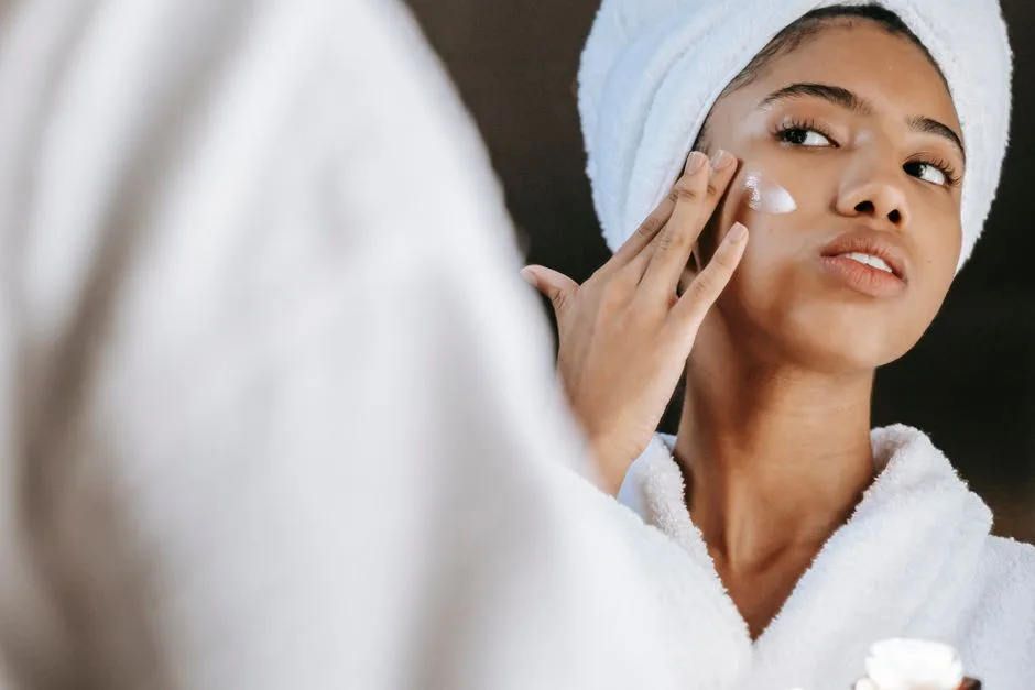 Low angle of ethnic female in towel looking at mirror while treating skin with moisturizing lotion during morning routine