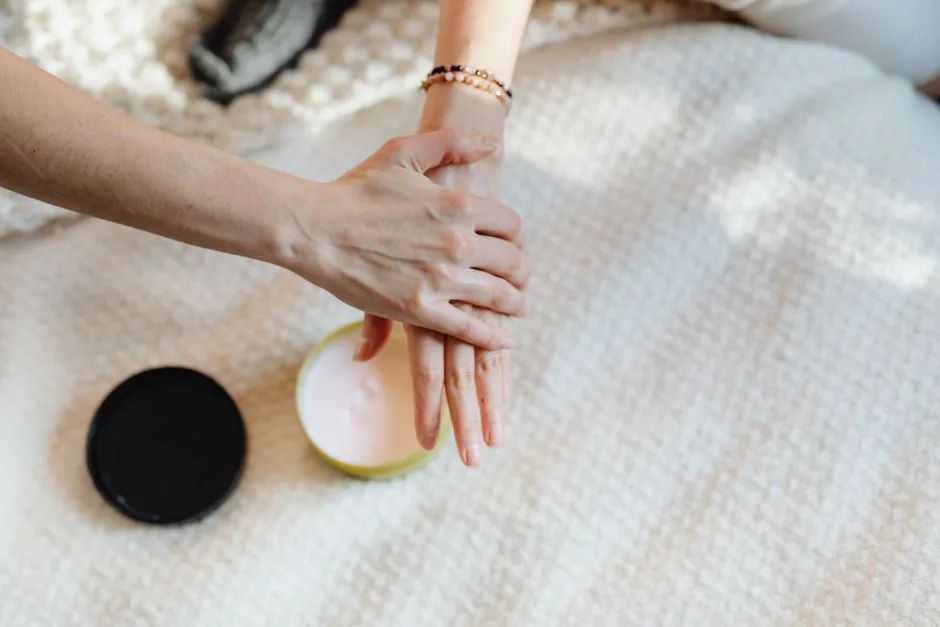A close-up of a woman applying moisturizer on her hands with a cream container on a cozy bed.