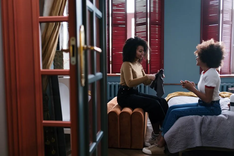 Two friends sitting indoors exchanging clothes, expressing camaraderie and style.