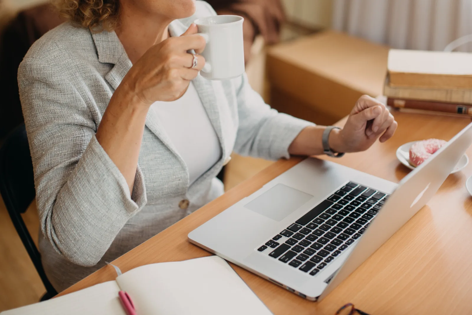 A woman in a light grey blazer sips coffee while engaged in work on her laptop. The workspace features books and a small plate of pastries. Sunlight fills the cozy atmosphere.