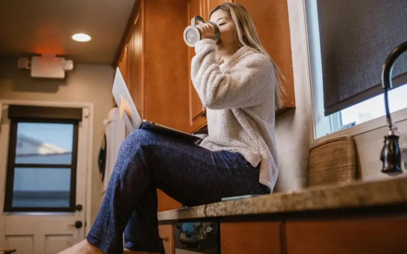 Casual morning scene of a woman enjoying coffee and using a laptop sitting on a kitchen counter.