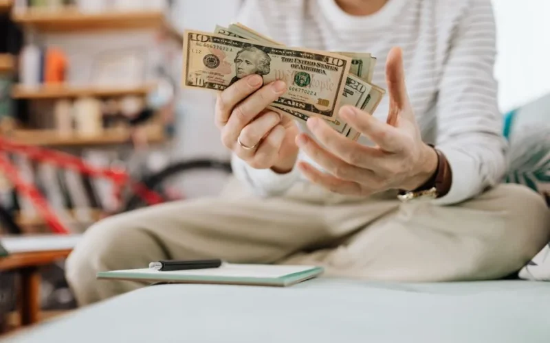 Man wearing casual clothes counts dollar bills while sitting on a sofa indoors.
