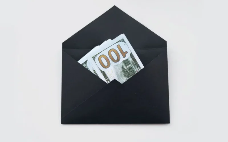 Hundred dollar bills partially visible in an open black envelope on a white background.