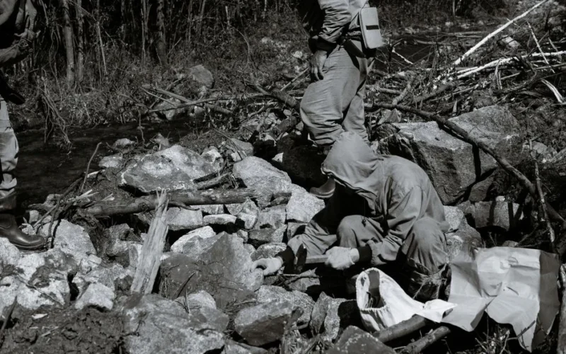 Black and white of anonymous male scientists with hammer among rough rocky formations against woods