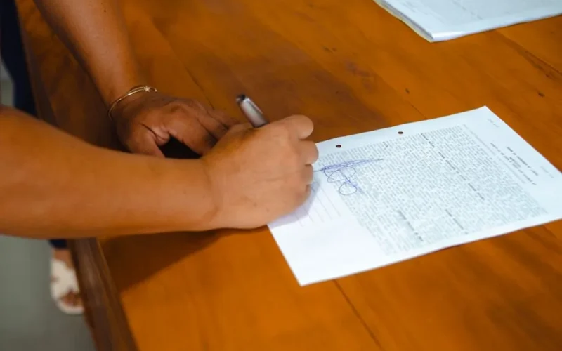 Close-up of hands signing a legal document on a wooden table indoors.