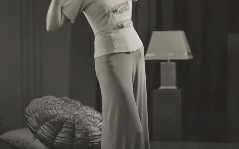 Woman posing in studio setting, leaning back to left, with raised arms and hands near her shoulders. In background a low bench with two cushions resting on it at left. A table lamp in centre and to right of woman.