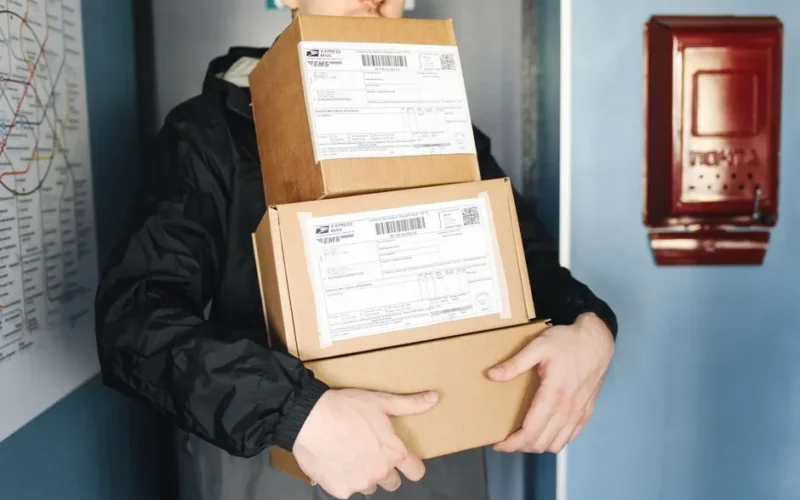 Courier carrying stacked cardboard packages for delivery service indoors.