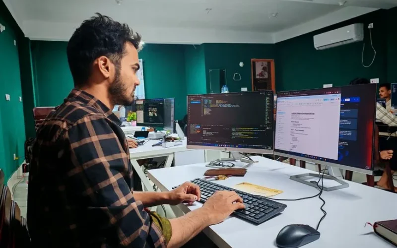 A software developer working on code at a dual monitor setup in a modern office.