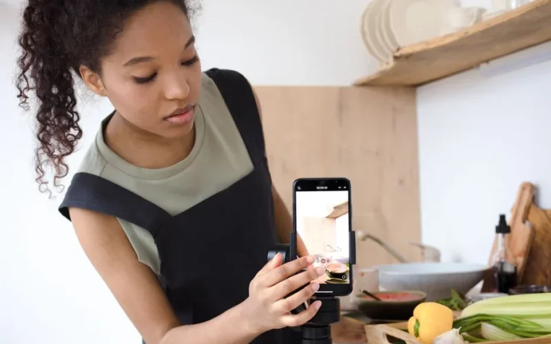 A young woman filming a cooking tutorial in her kitchen using a smartphone.