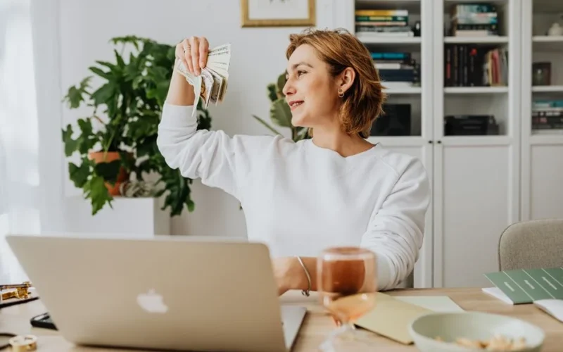 Smiling woman holding cash, working at home office with a laptop, symbolizing financial success.