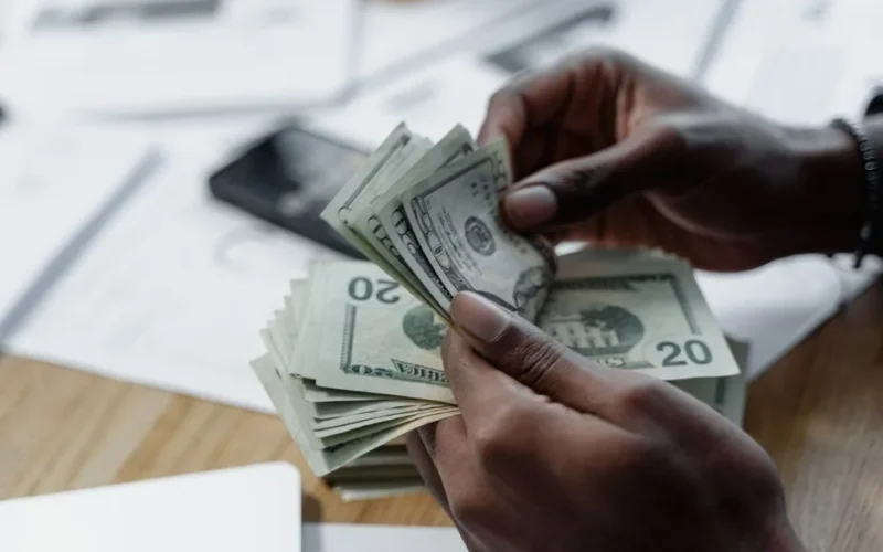 A close-up of hands counting US dollar bills on a wooden table with documents and a smartphone nearby.