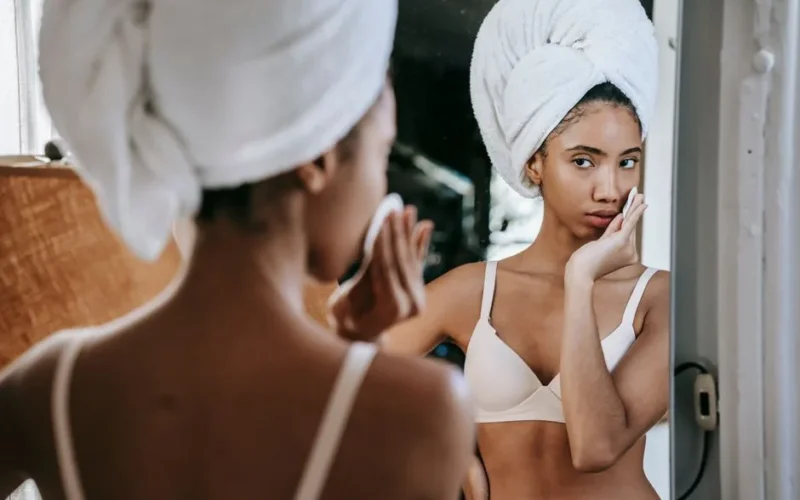 Serious ethnic female looking at mirror while moisturizing skin with lotion during daily beauty routine in bathroom