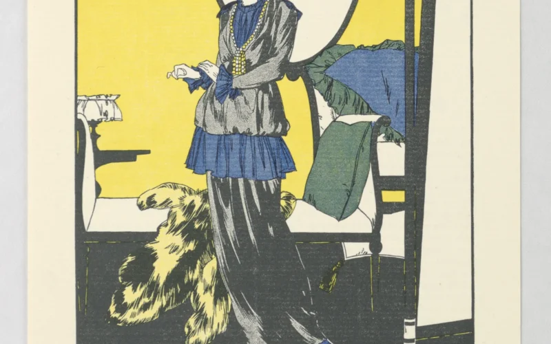 In an interior, a woman in matching suit jacket and floor-length skirt, looks at self wearning a large black hat in the standing mirror to her left. Background, fur coat sits upon day bed behind her.