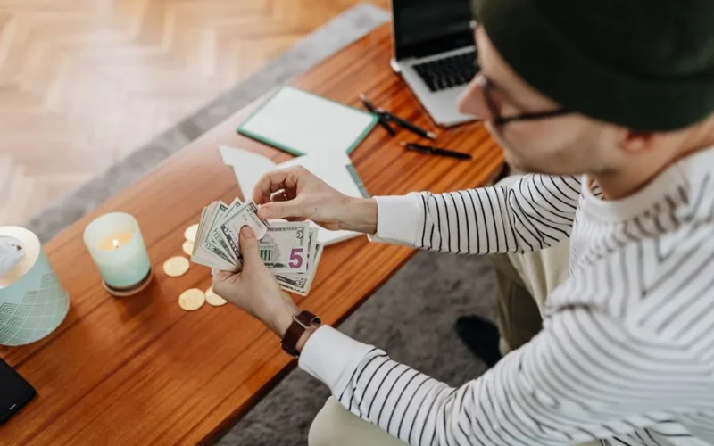 A person counts US dollars at a wooden table with a laptop, candle, and coins in view, creating a cozy atmosphere.