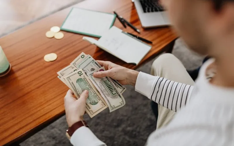 A person counts US dollar bills with a notebook and coins on the desk.