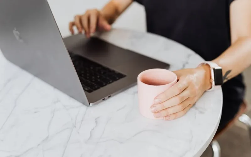 A person working on a laptop while holding a coffee cup at a marble table.