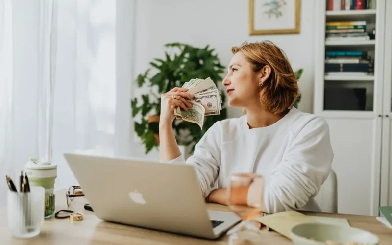 Woman sitting at desk holding cash while working on a laptop in a cozy home office setting.