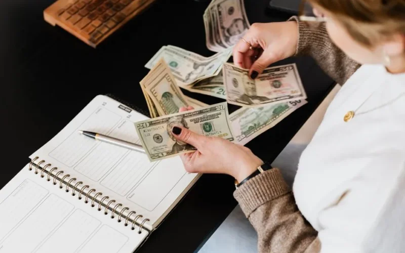 Woman organizing finances, counting dollar bills at desk with open notebook.