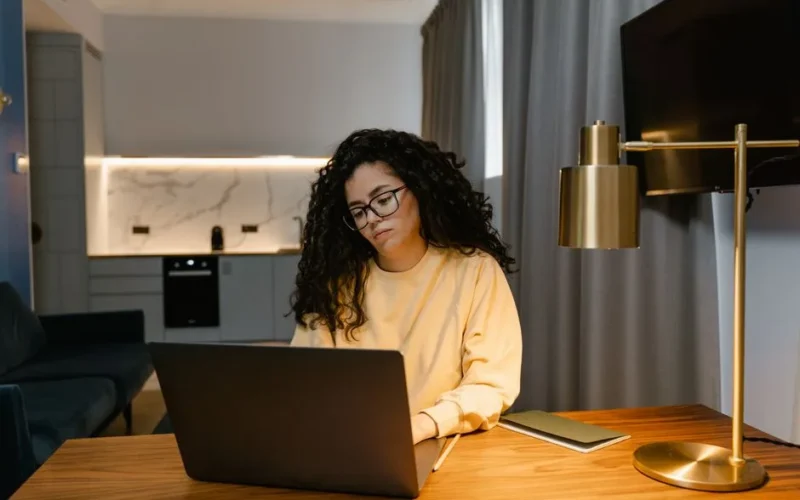 Woman in a yellow sweatshirt using a laptop at home, focusing on remote work in a cozy, modern interior.