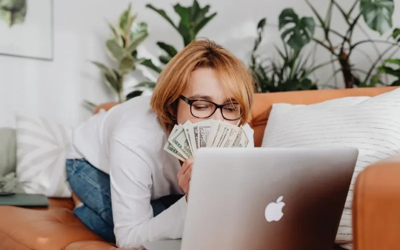 Woman holding cash while using a laptop on a cozy sofa at home, conveying financial success.