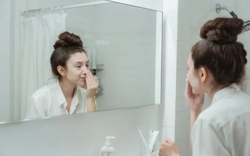 Caucasian woman in bathroom applying skincare product while looking in the mirror.
