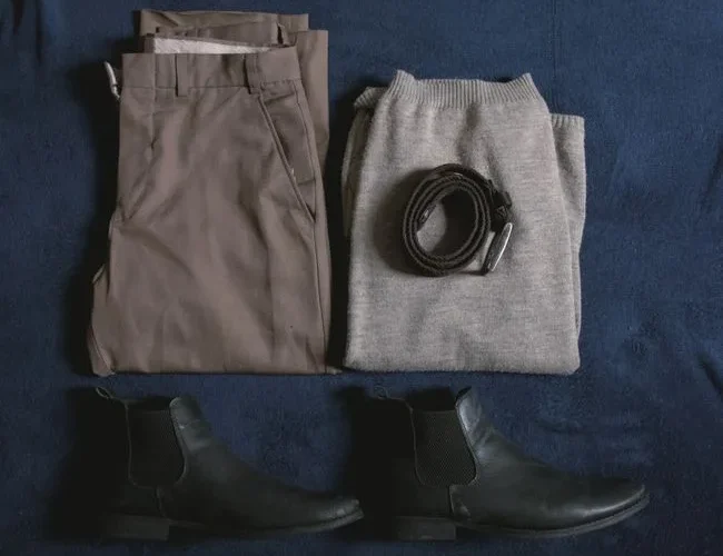 Men's clothing ensemble including trousers, sweater, boots, and belt arranged on a blue surface.
