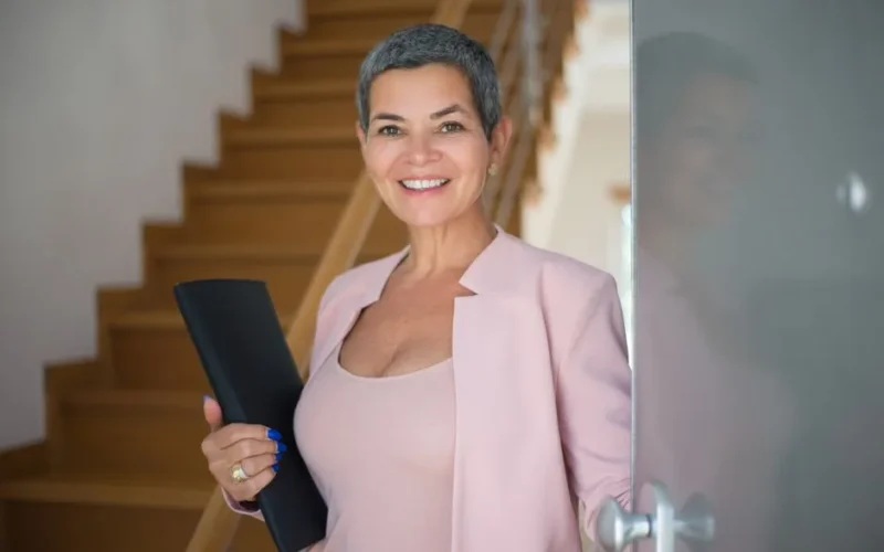 Confident senior woman entering a modern office setting, holding documents and smiling.