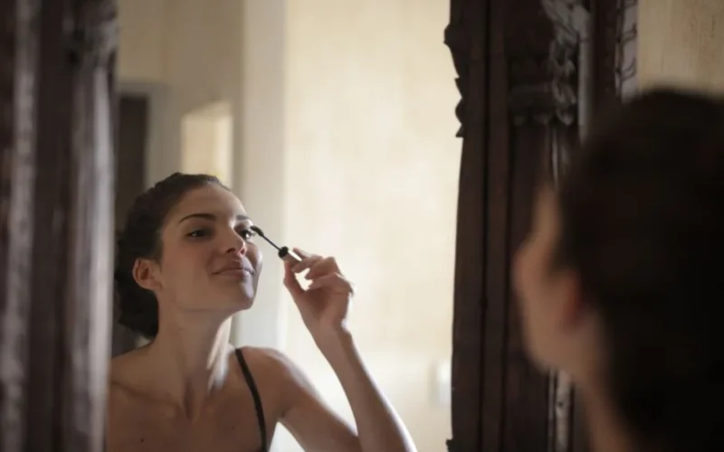 A woman applies mascara while looking in a stylish wooden mirror indoors.