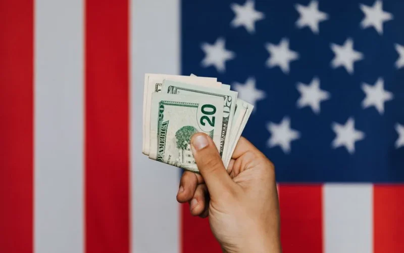 Unrecognizable male demonstrating dollars before official flag of USA on background