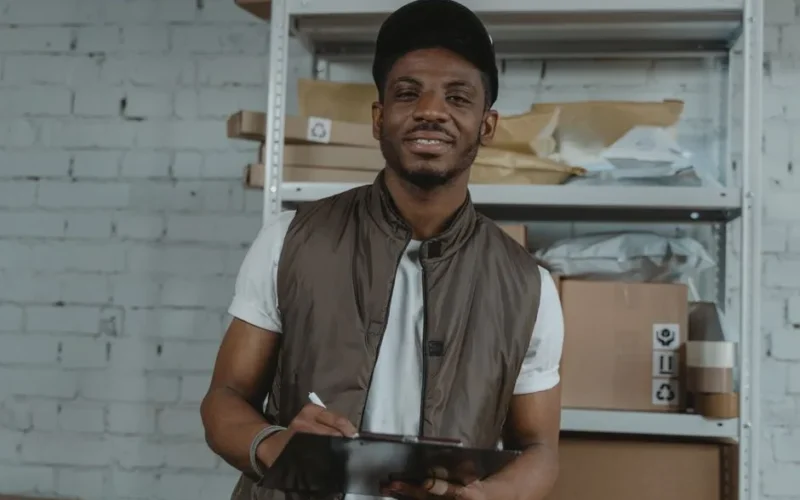 Warehouse employee holding a clipboard, surrounded by packages and smiling.