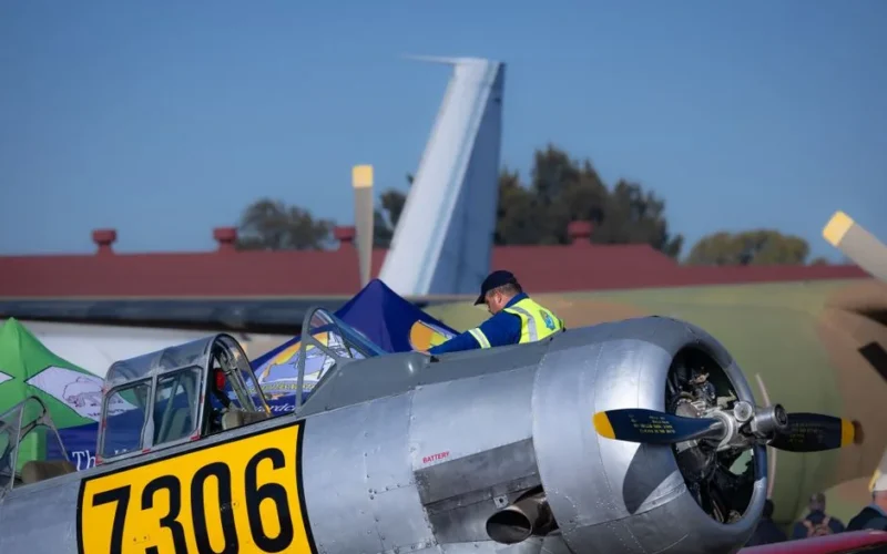 A mechanic inspecting and maintaining a vintage aircraft outdoors at an airfield during the day.