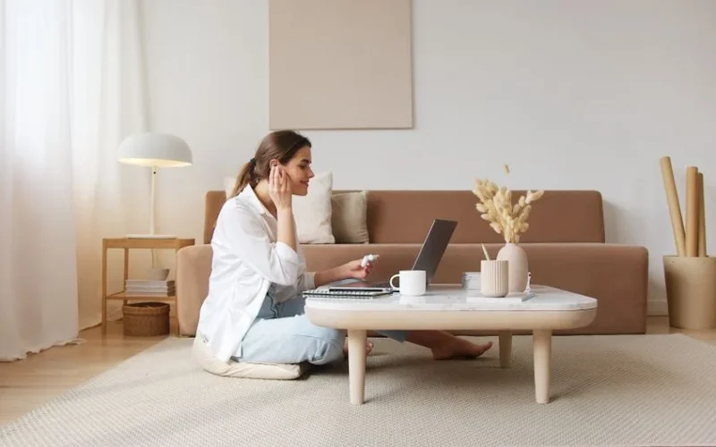 Side view full body young positive female in casual clothes using wireless earbuds while having conversation via video call on laptop and sitting on floor in cozy apartment