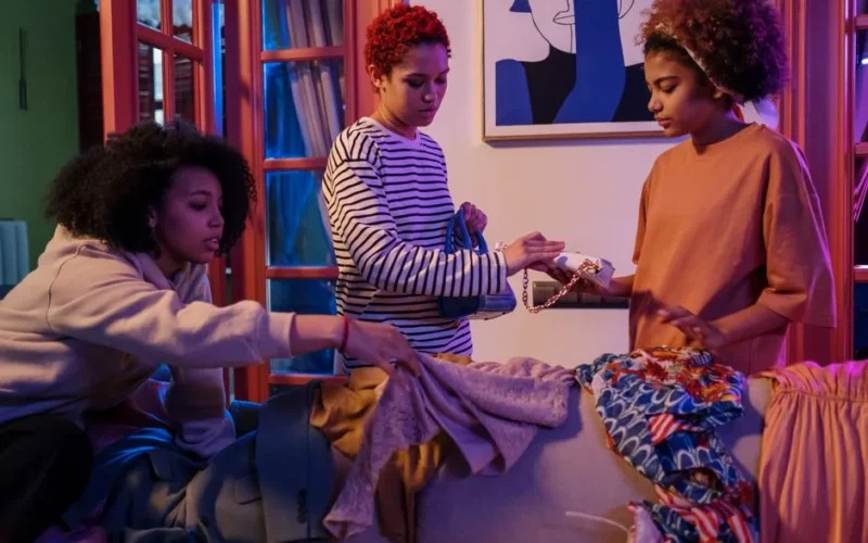 Three women in cozy setting swapping clothes and accessories indoors.