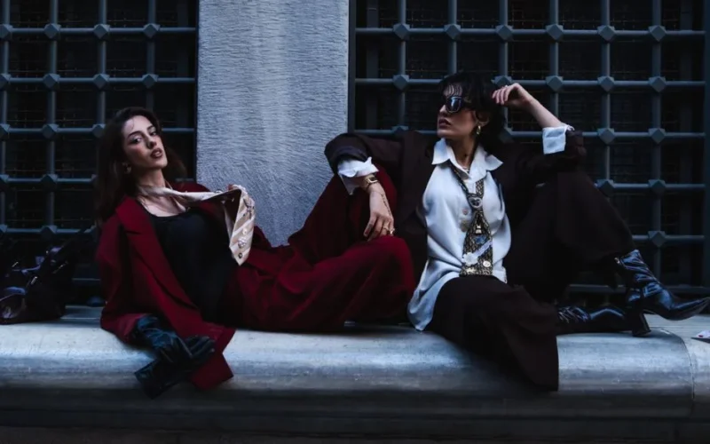 Two fashionable women in stylish outfits posing against an urban backdrop.