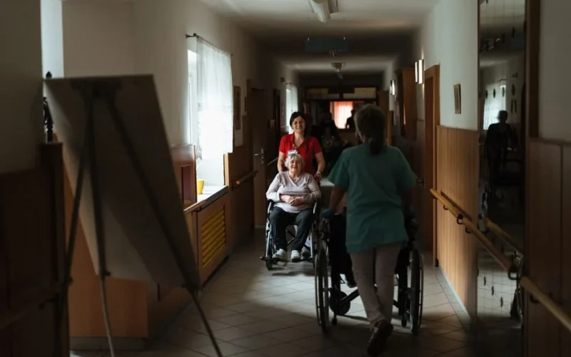 Caregiver assisting elderly woman in a wheelchair through a retirement home corridor.