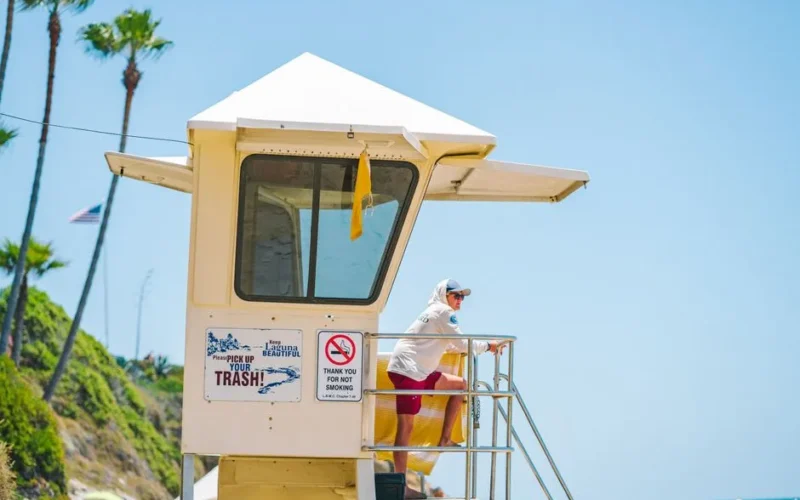 A lifeguard wearing a cap and hoodie watches over a sunny beach from a tower.