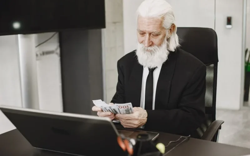 Senior businessman in suit counting money at office desk with a laptop. Elegance and financial focus in modern workspace.