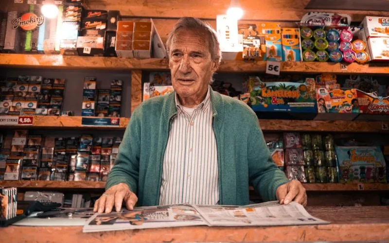 An elderly shopkeeper stands behind the counter in a Lisbon newsstand, surrounded by colorful products.