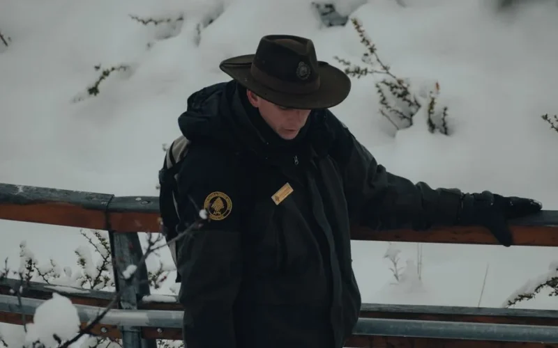 A park ranger in winter attire stands on a snow-covered bridge, reflecting tranquility in nature.