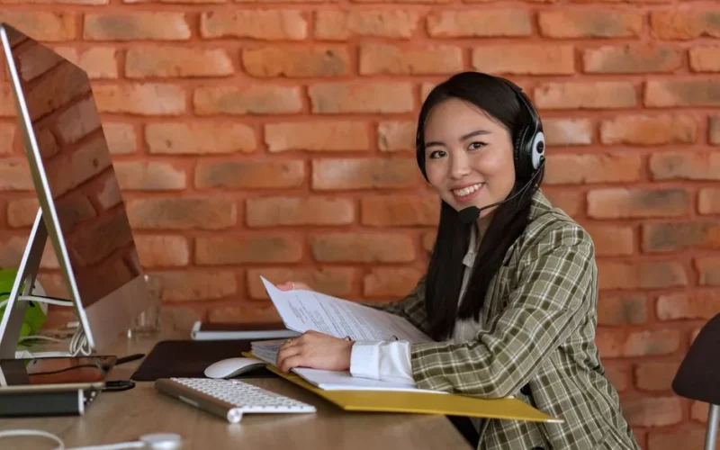 A woman working as a call center agent sitting at a desk with a computer and files.