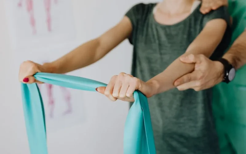 Crop unrecognizable orthopedist in uniform and wristwatch supporting shoulder and reached arm of faceless female patient with resistance band in doctor office