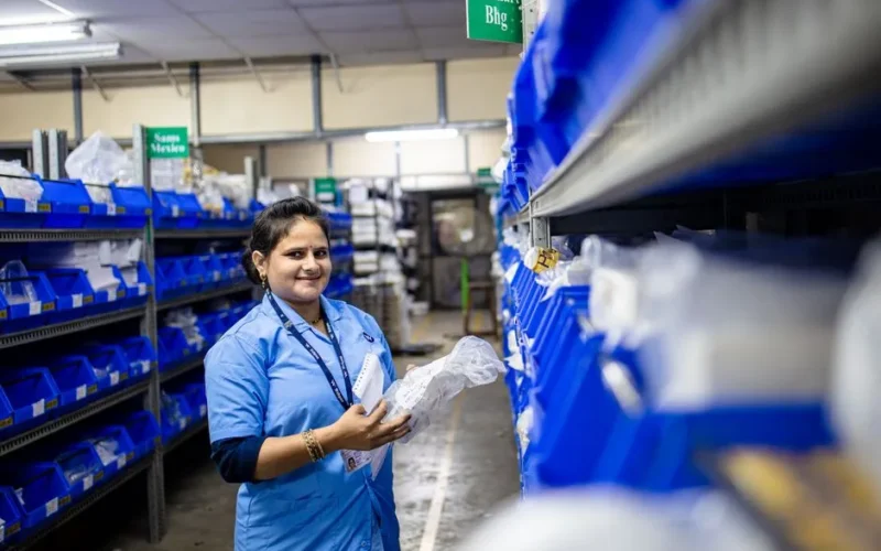 Smiling woman in a warehouse organizing inventory on shelves with blue bins.